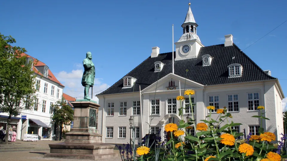 Billede af Randers: Statue på torv med hvid bygning og klokketårn bag gule blomster i forgrunden.