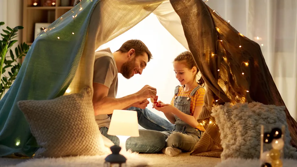 Father and daughter exchanging a gift inside a blanket fort with string lights.