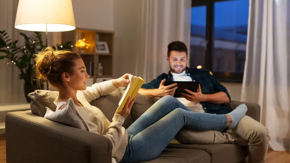 Couple on couch, woman reading book and man using tablet.