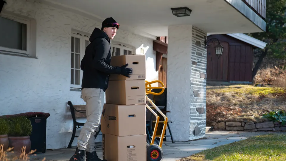 Leveringsarbeider stapper pappesker på håndtralle utenfor hus.