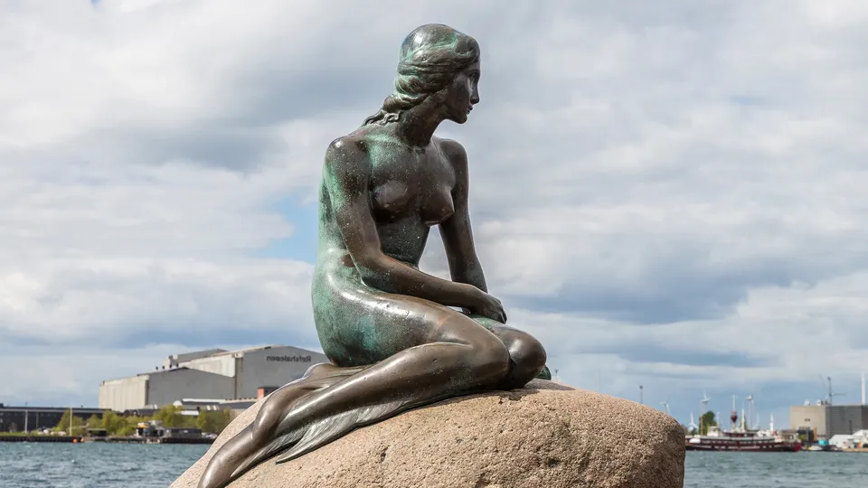 Bronsestatue av en sittende havfrue på en stein ved vannet.