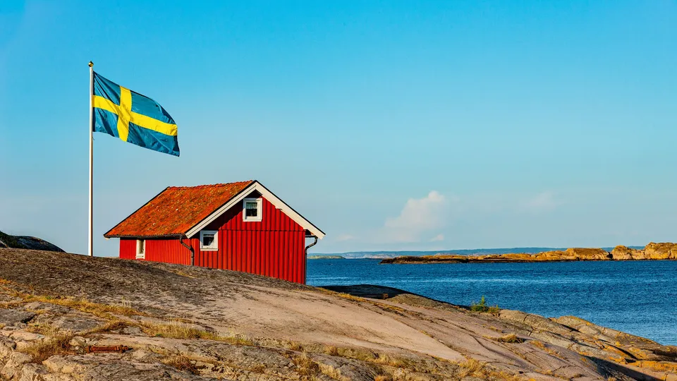 Svensk flagg som vaier over rød hytte på steinete kystlinje.