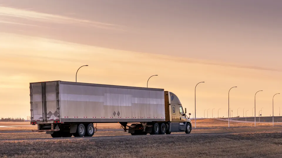 Lastebil med stor lastetrailer kjører på tom motorvei ved solnedgang.