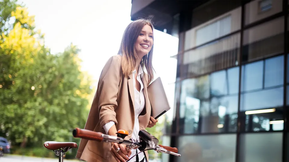 Mujer sonriente sosteniendo una bicicleta con portátil, de pie frente a un edificio moderno.