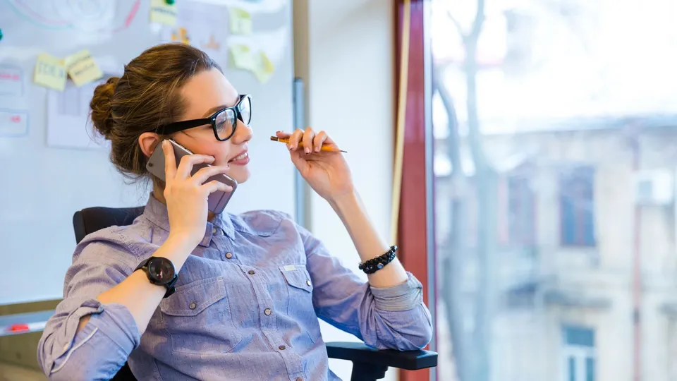 Kvinde taler i telefon og holder blyant ved skrivebord med whiteboard bag hende.