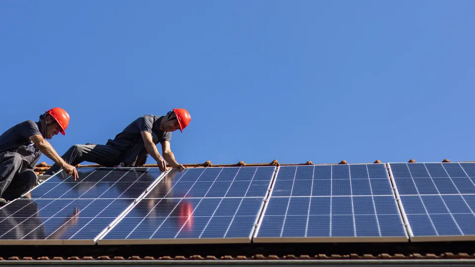 Dois trabalhadores com capacetes a instalar painéis solares num telhado.