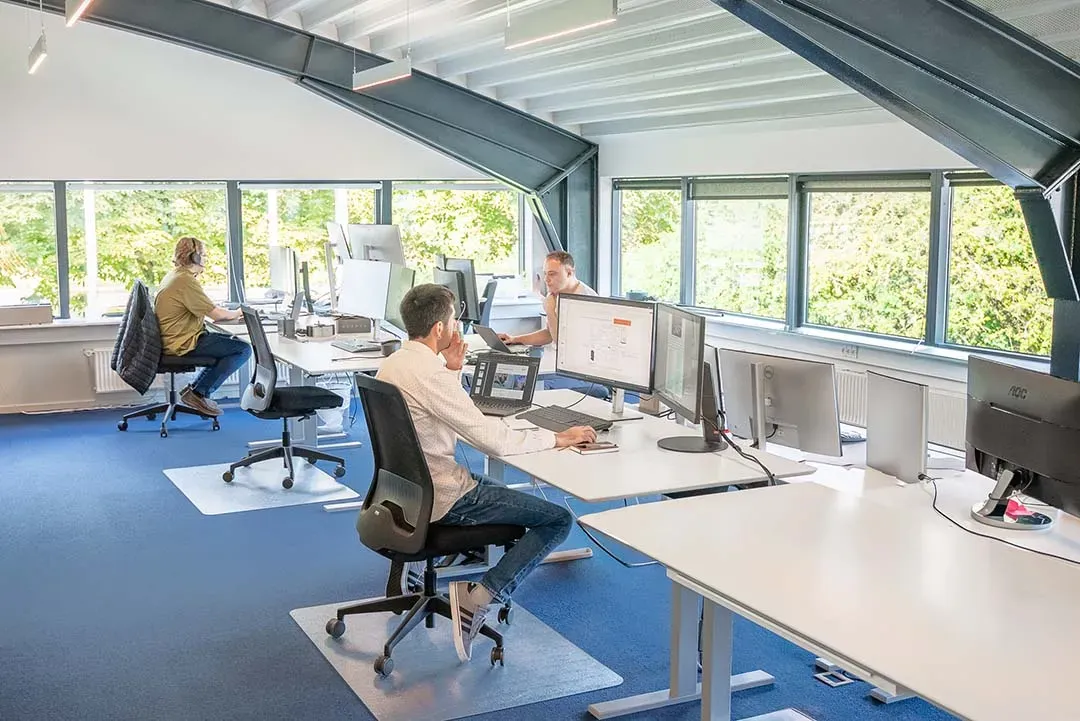 Tre personer arbejder ved computerborde på et lyst kontor med store vinduer.