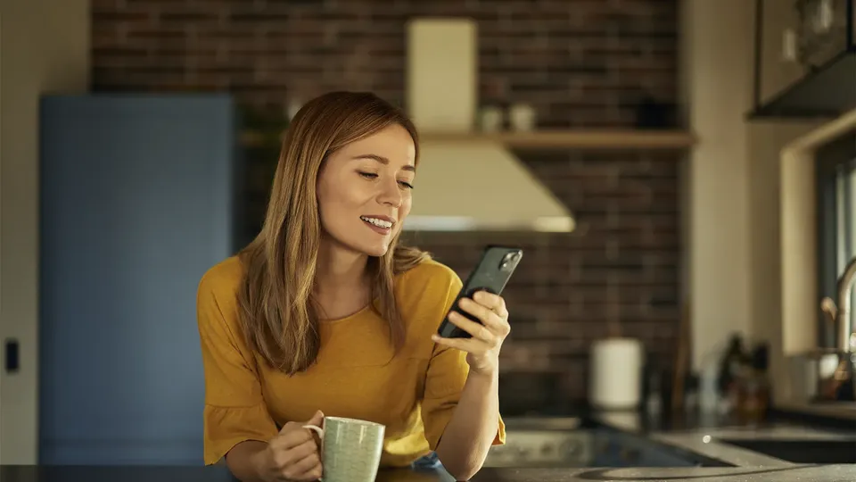 Kvinde i gul skjorte holder telefon og kop i hånden i hendes køkken og smiler til skærmen.