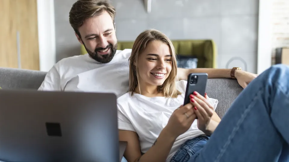Par på sofa smiler mens de ser på smartphone og laptop.