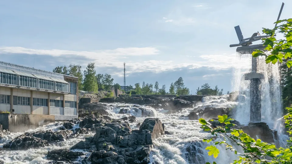 Powerful white-water rapids tumbling over dark rocks beside an industrial building, with a metal sculpture on the right and spray falling over it, framed by bright green maple leaves under a pale blue sky.