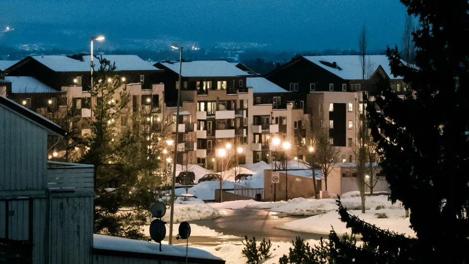 Snødekte leilighetsbygg belyst i skumringen med gatelys og opplyste vinduer.