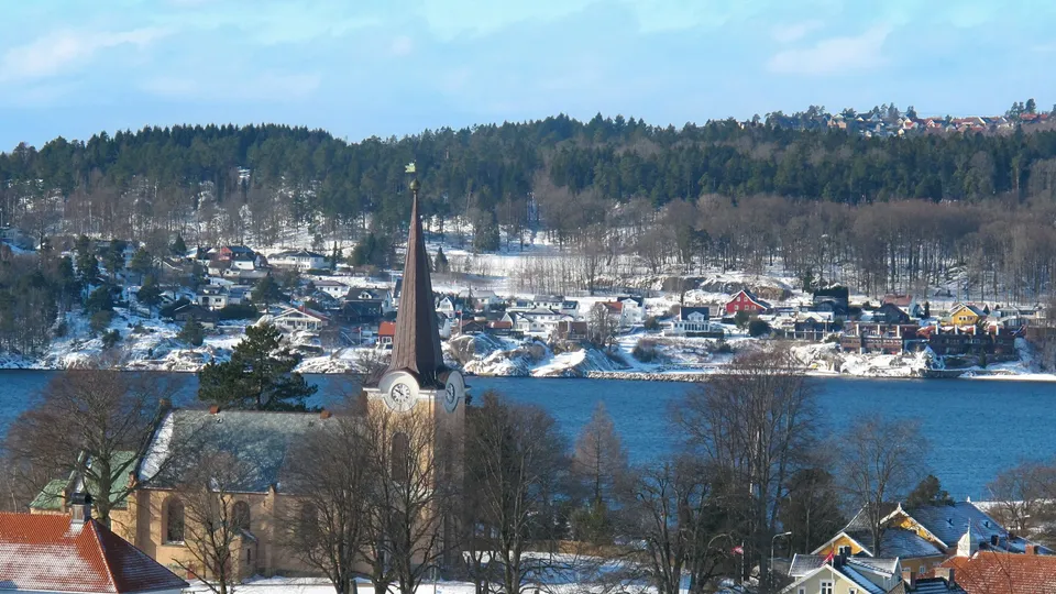 Kirke med høy spir utsikter over snøkledd kystby over blå fjord.