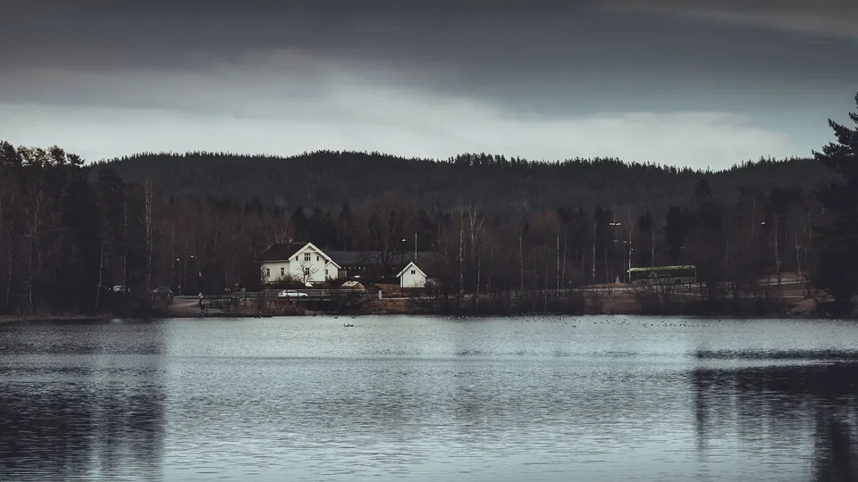 Hvitt hus og uthus på andre siden av stille innsjø med skogsbackdrop.