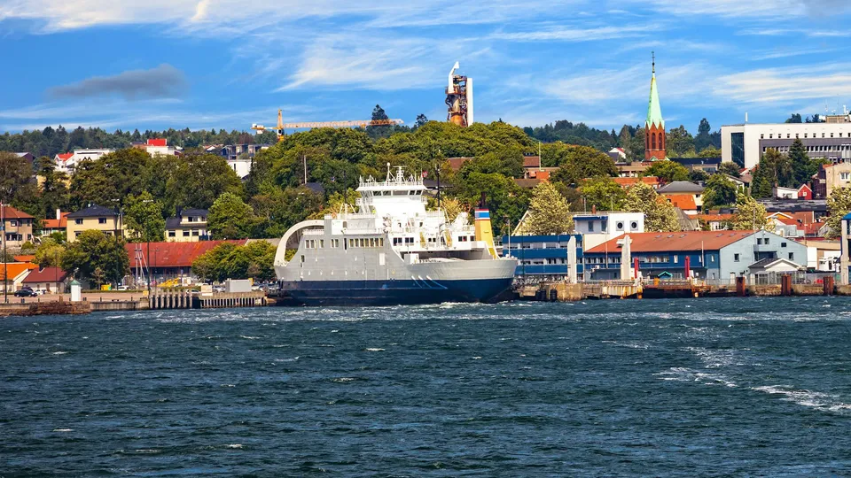 Hvit og blå ferge fortøyd ved kai med fargerike bygninger ved vannkanten og kirketårn i bakgrunnen.