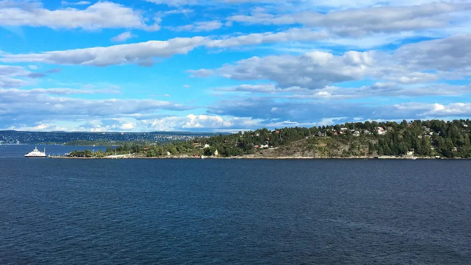 Hvit passasjerferry krysser blå fjord mot skogskledd odde med hus.