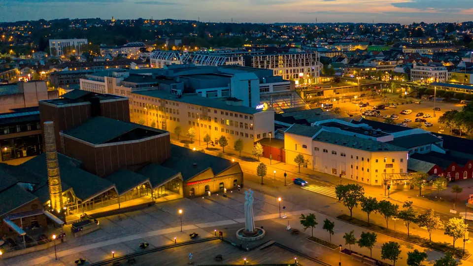 Aerial view of Sandnes city centre at dusk, with warmly lit streets and buildings, a brick cultural venue with a tall chimney, a white sculpture on a waterfront plaza, and a pink-and-blue evening sky above the surrounding residential hills.