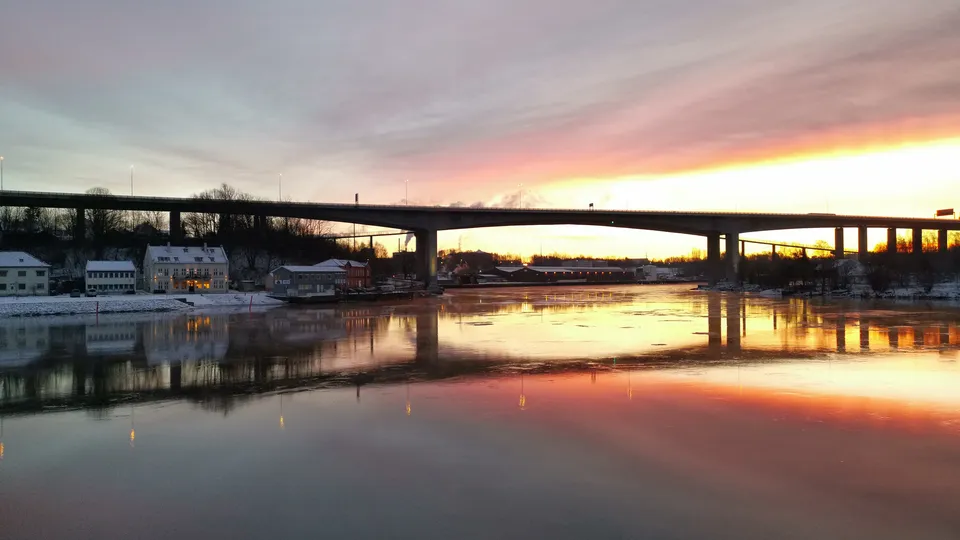 Bro som spenner over rolig elv ved solnedgang med bygninger ved vannkanten og fargerik himmelrefleksjon.