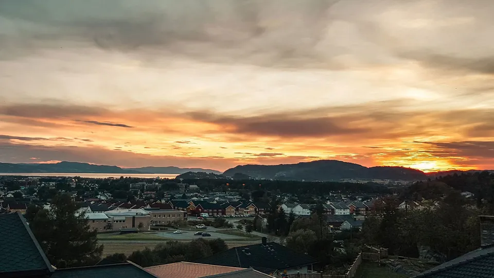 A vivid orange and golden sunset over a residential area, with houses and trees on the hillside, a distant fjord and dark silhouetted hills on the horizon.