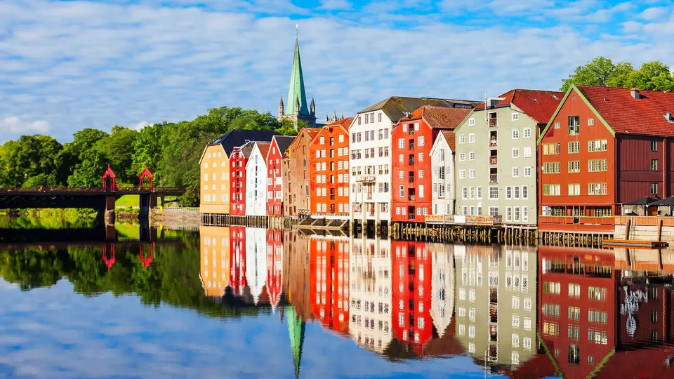 Fargerike bygninger i Trondheim ved vannkanten reflektert i stille vann med Nidarosdomen i bakgrunnen.