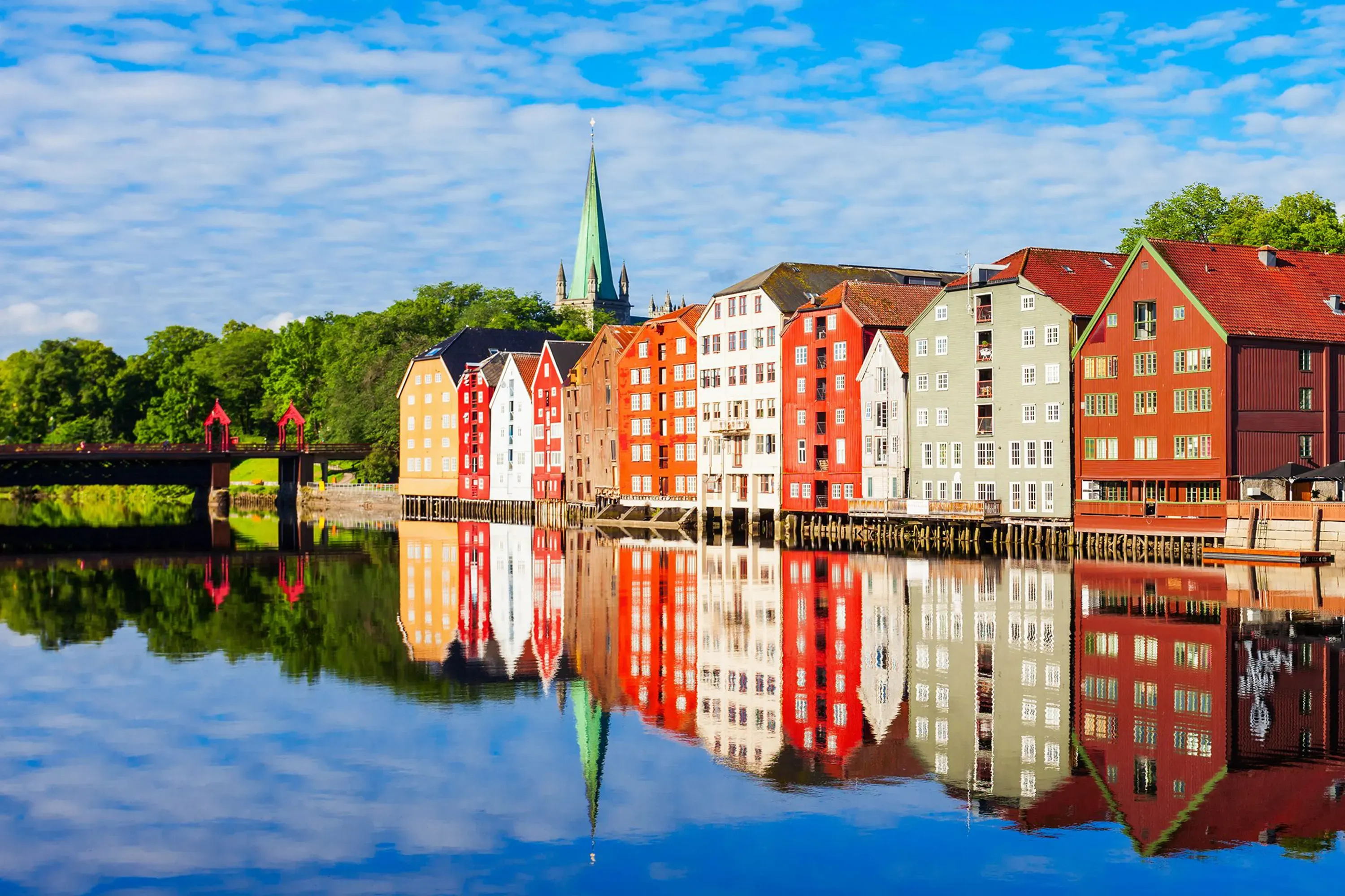 Fargerike bygninger i Trondheim ved vannkanten reflektert i stille vann med Nidarosdomen i bakgrunnen.