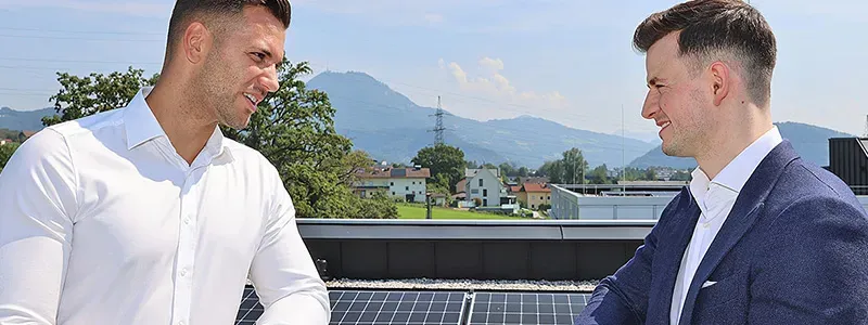 ENKO GmbH – zwei Geschäftspartner im Gespräch auf Dachterrasse mit Photovoltaikanlage vor alpiner Kulisse