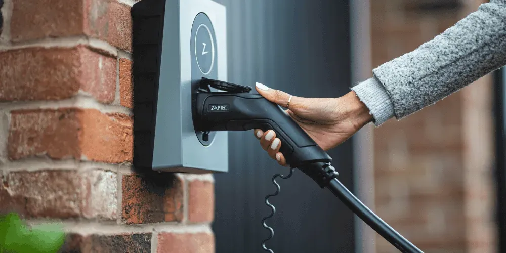 Woman plugging a Zaptec home EV charger into a wall-mounted unit on a brick exterior wall
