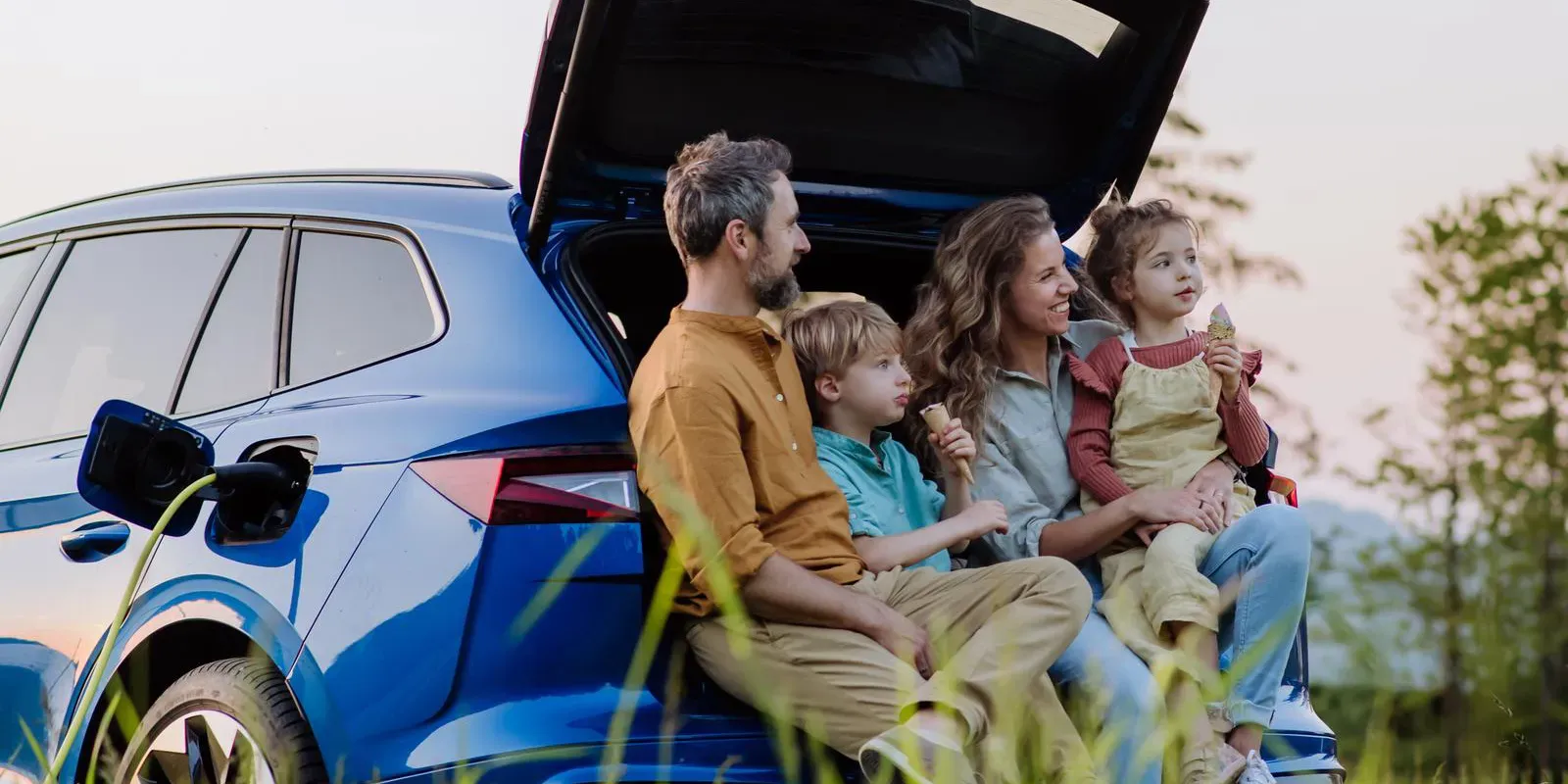 Famiglia di cinque persone seduta nel bagagliaio aperto di un'auto elettrica blu in carica