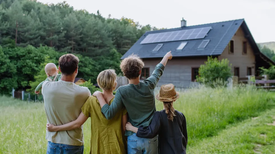 Eine fünfköpfige Familie steht auf einem Feld und blickt auf ein Haus mit Solarmodulen.