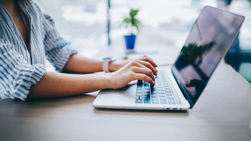 Person skriver på bærbar computer ved skrivebord med lille pottet plante og vindue i baggrunden.