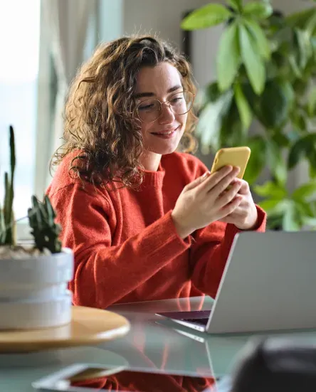 Mujer sosteniendo un smartphone mientras trabaja en una laptop rodeada de plantas.