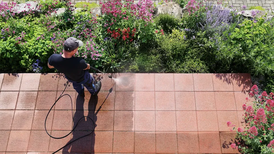 Person vasker en flisebelagt terrasse med højtryksrenser omgivet af blomstrende planter.