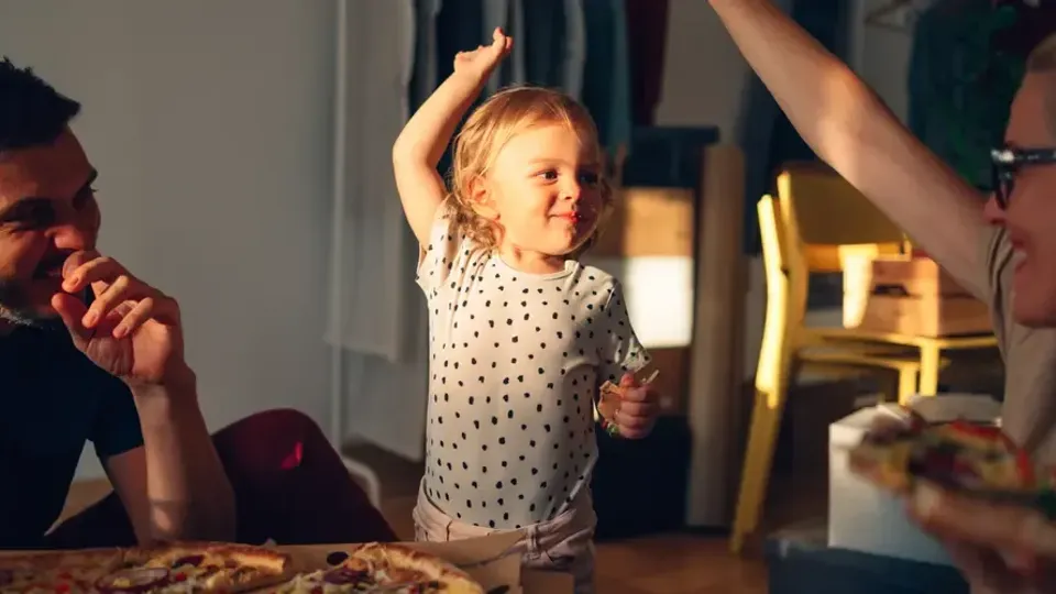 Litet barn dansar med armarna höjda medan vuxna tittar vid bordet.