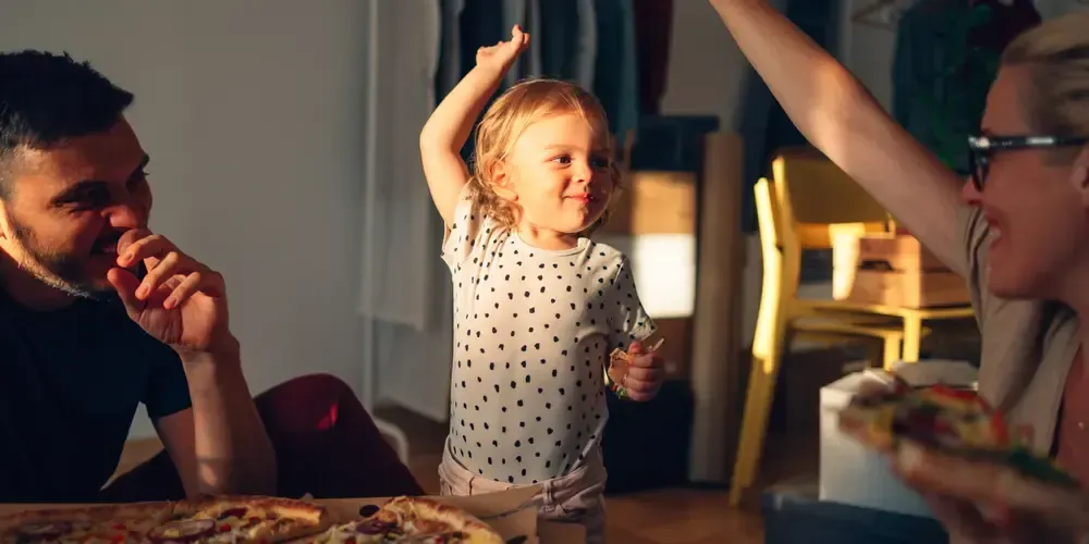 Litet barn dansar med armarna höjda medan vuxna tittar vid bordet.