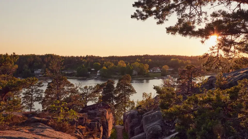 Solnedgång över skärgårdslandskap med klippor, tallar och stilla vatten sett från höjd.