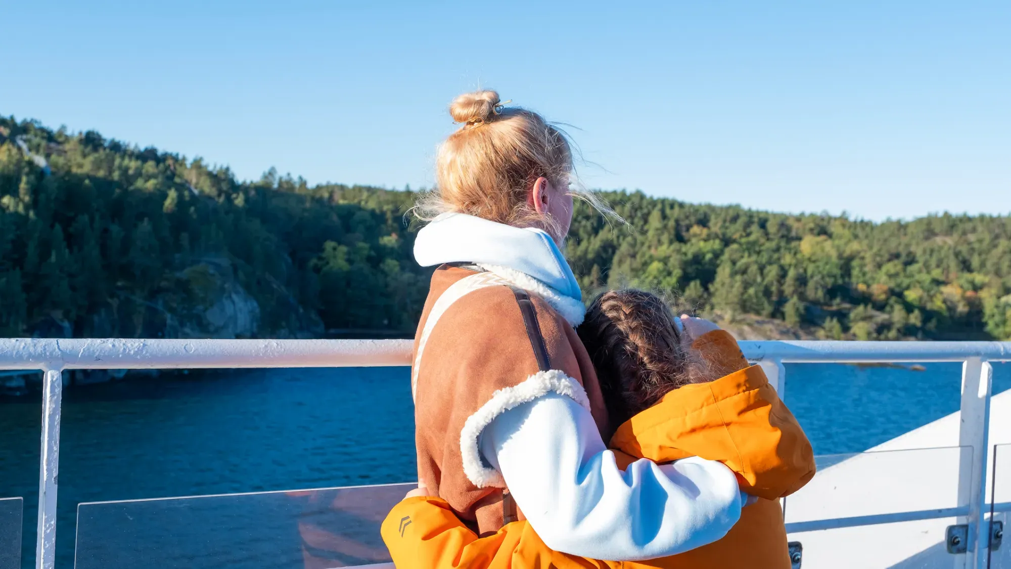 Kvinna och barn i orange jacka står på färjedäck och tittar ut över blått hav och grön skärgård i solsken.