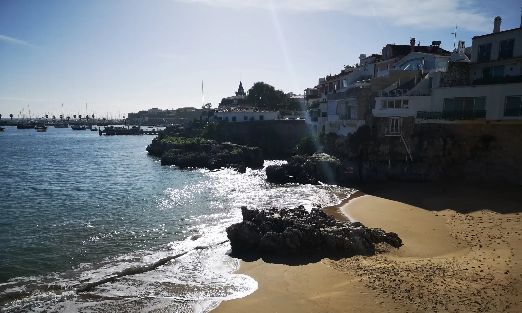 Strand och klippor vid havet i Cascais med bostadshus och en småbåtshamn längs kusten.
