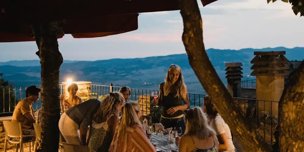 Grupp människor äter middag utomhus på en terrass med utsikt över kullriga kullar.