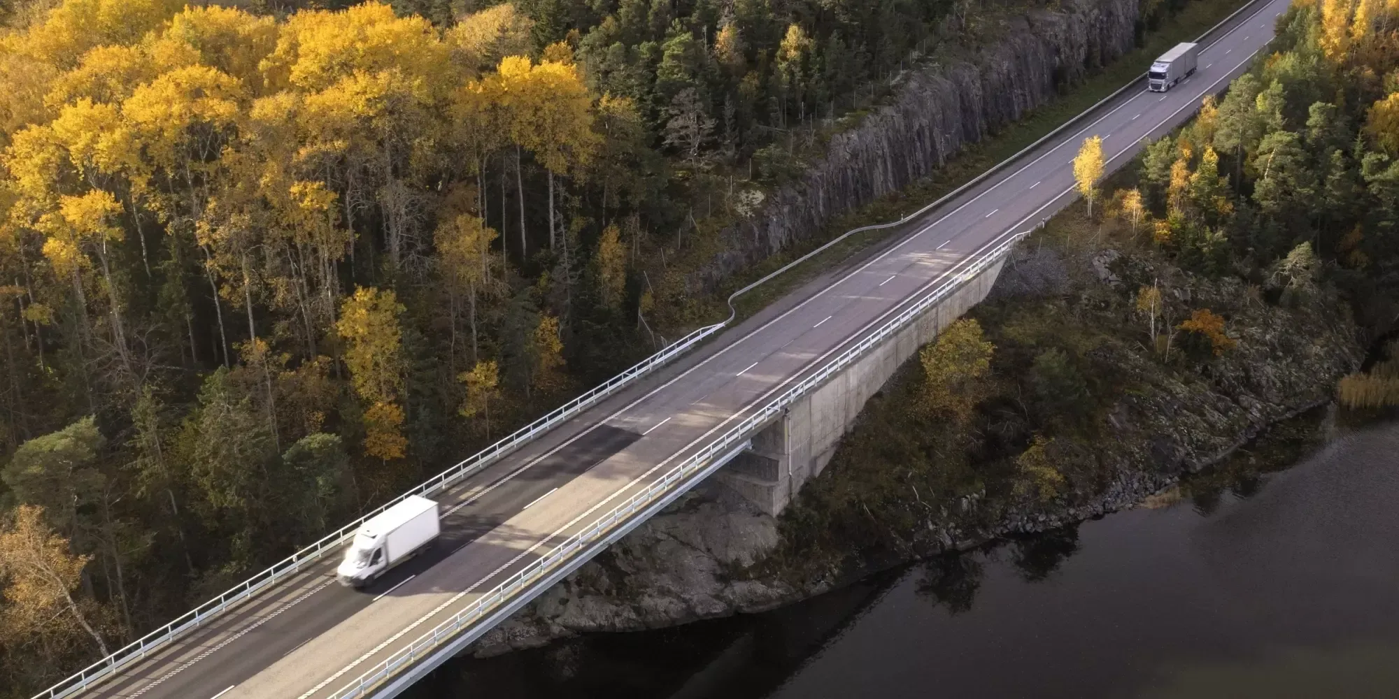 En motorväg ovanifrån med höstig skog runtomkring, och två lastbilar på vägen.