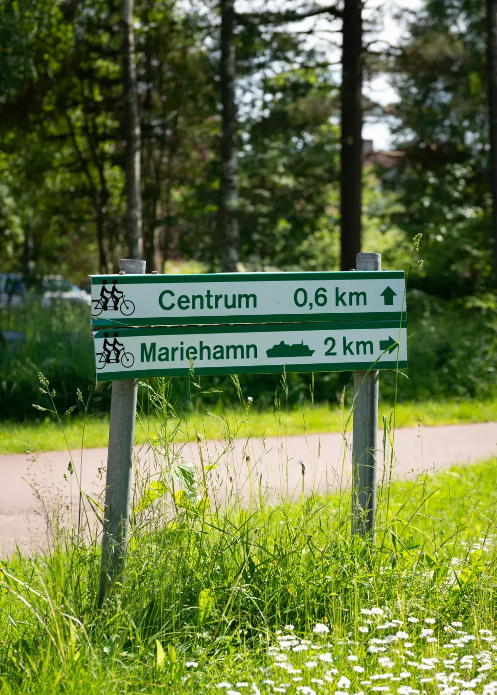 Grön vägskylt vid cykelväg som visar riktning mot Centrum 0,6 km och Mariehamn 2 km, omgiven av träd och grönska.