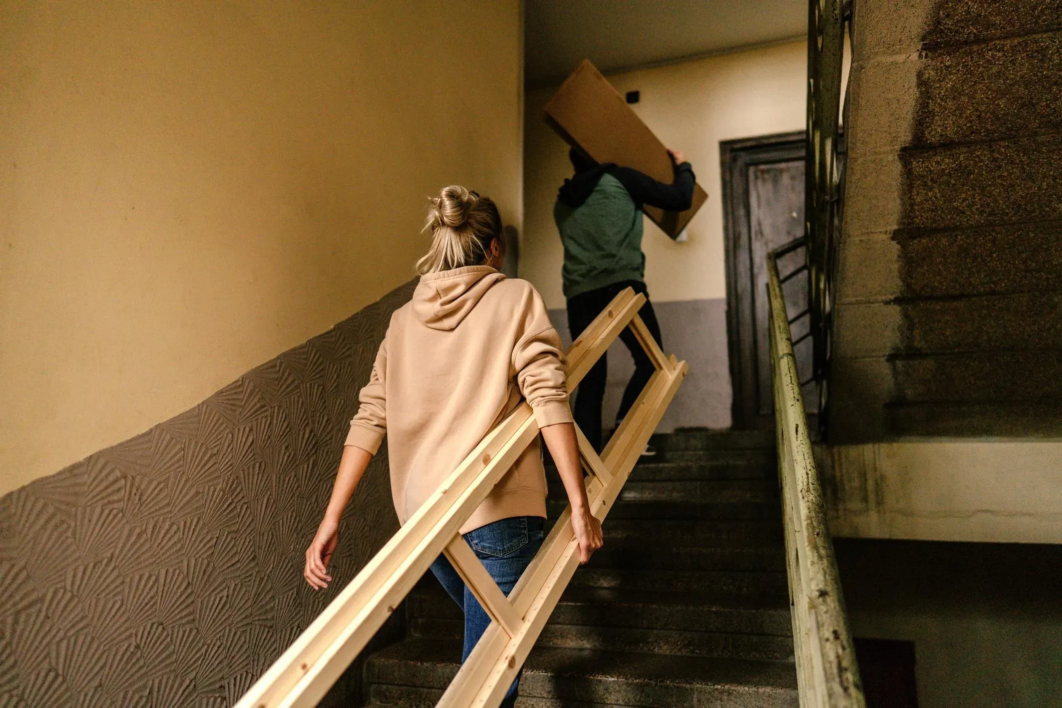 Två personer bär en trästege och en flyttkartong upp för en trappa under en flytt.