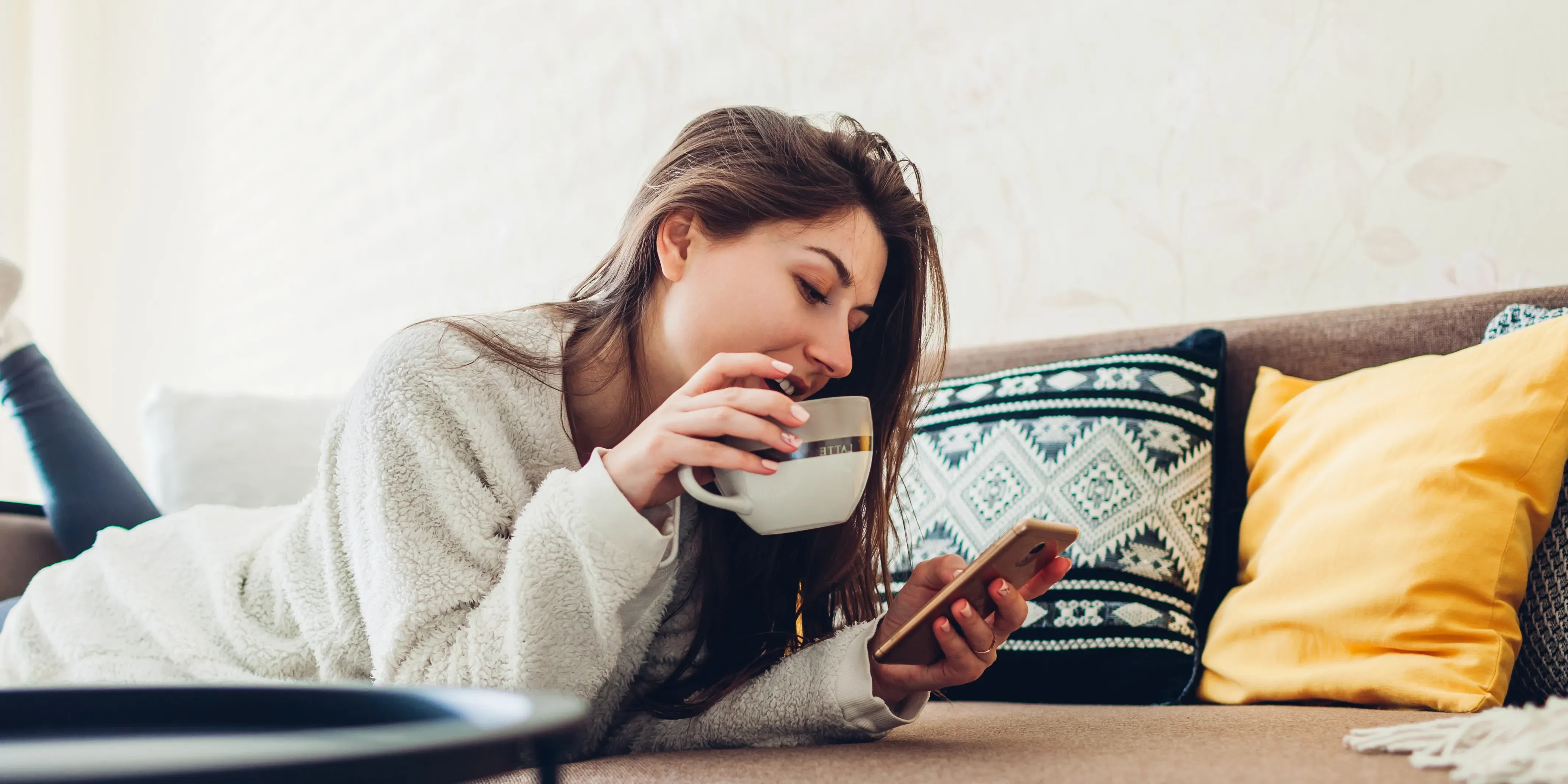 Kvinne på sofa som holder kaffekkopp og ser på telefonen.