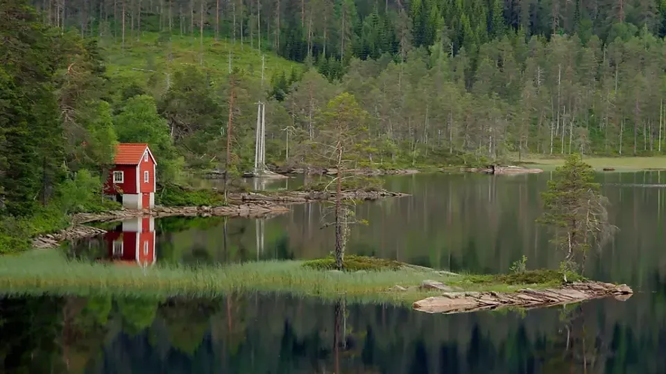 Röd stuga med vit skorsten reflekterad i lugn sjö omgiven av skog.
