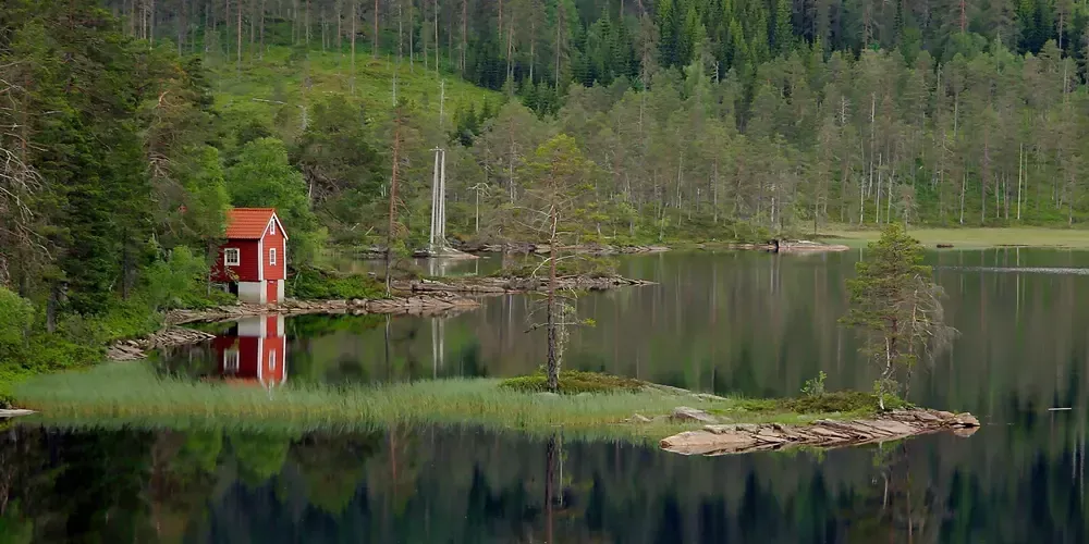 Röd stuga med vit skorsten reflekterad i lugn sjö omgiven av skog.