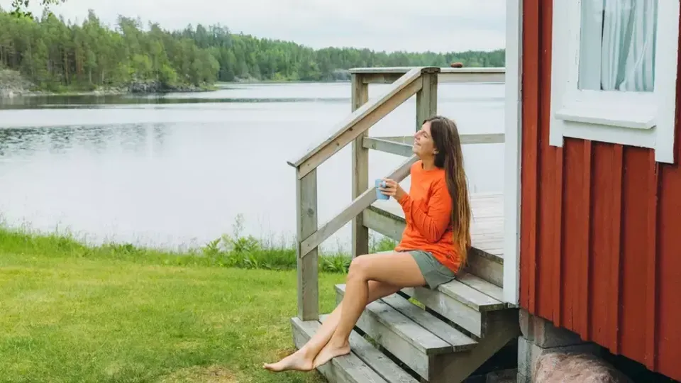 En kvinna dricker en kopp kaffe på trappen till sitt svenska röda fritidshus.