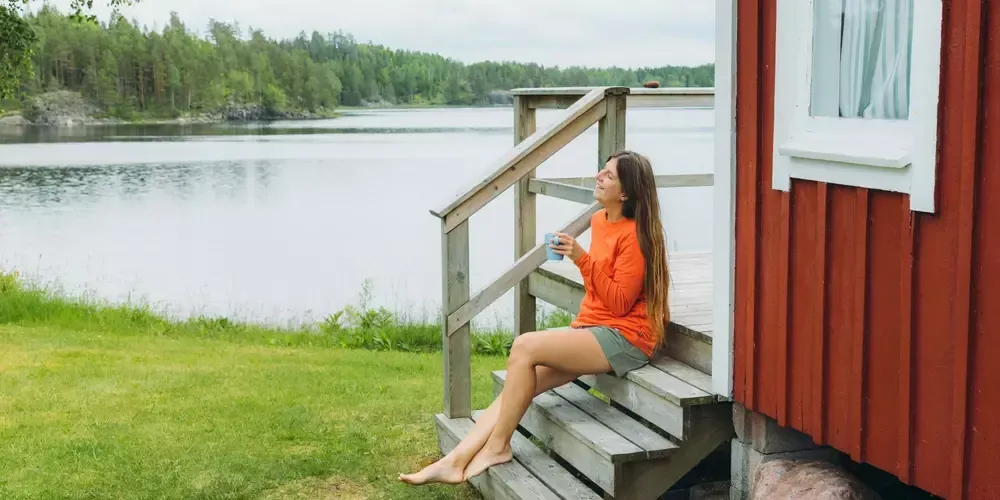 En kvinna dricker en kopp kaffe på trappen till sitt svenska röda fritidshus.