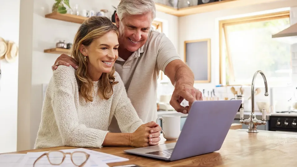 Hombre y mujer mayores sonriendo juntos frente a una laptop en la barra de la cocina.