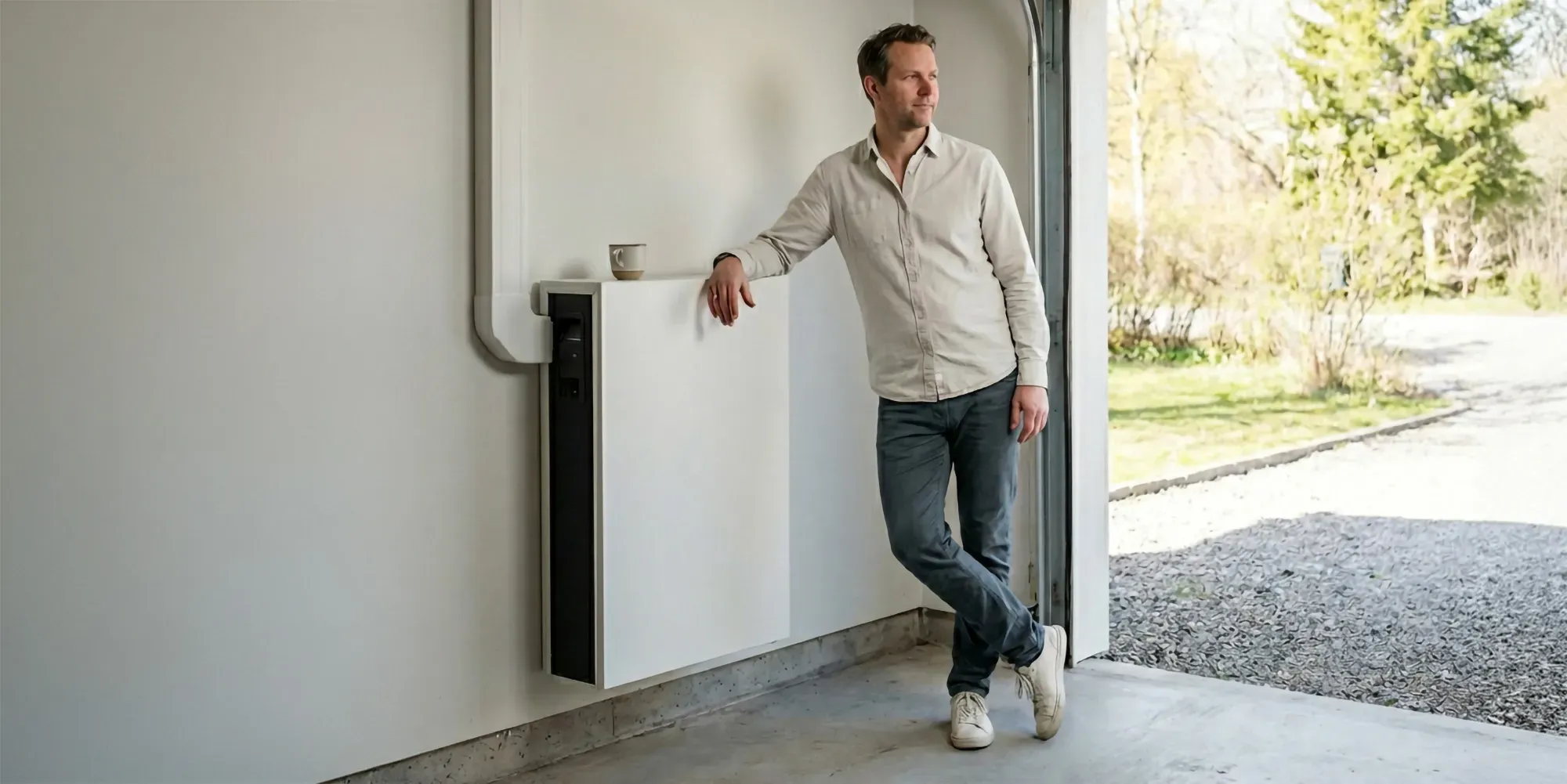 En stolt medelålders svensk man som lutar sig mot sitt nyinstallerade hembatteri i ett garage och ser nöjd ut. Han har ställt en kaffekopp på batteriet.