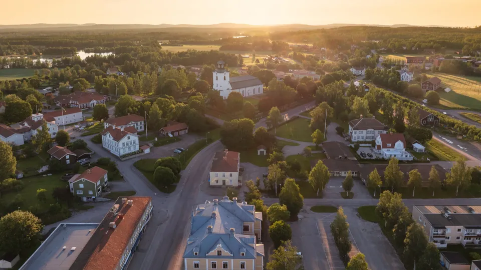 Flygfoto över Alfta i Ovanåker med en vit kyrka och bostadshus omgivna av skogar och åkrar vid solnedgång.