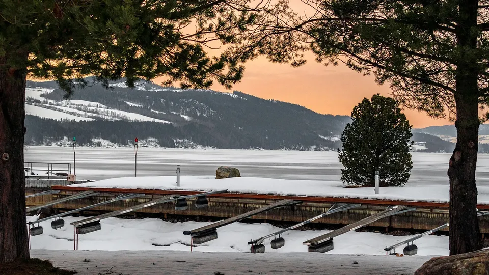 Frossen innsjø med snødekte fjell innrammet av furutrær og tom båtbrygge.