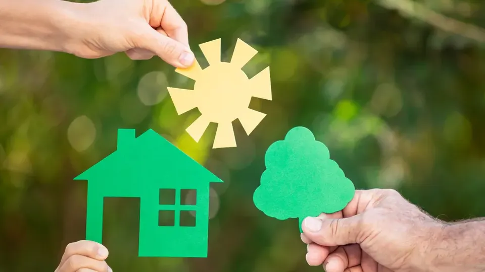 Due mani tengono ritagli di carta verde di una casa e un albero con un sole giallo sopra.